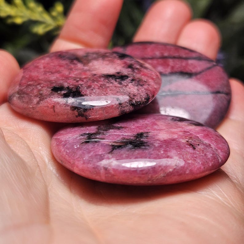 May include: Three polished, circular rhodonite stones featuring a vibrant pink base color with distinct black manganese oxide inclusions and veins. These smooth, flat, coin-shaped mineral specimens exhibit a glossy, reflective surface texture throughout.