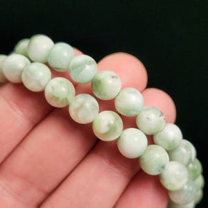 Peut inclure: Gros plan sur un bracelet compos&eacute; de perles rondes vertes claires et blanches. Les perles ont un aspect marbr&eacute;, avec des tourbillons de vert et de blanc. Le bracelet est tenu dans une main sur fond sombre.