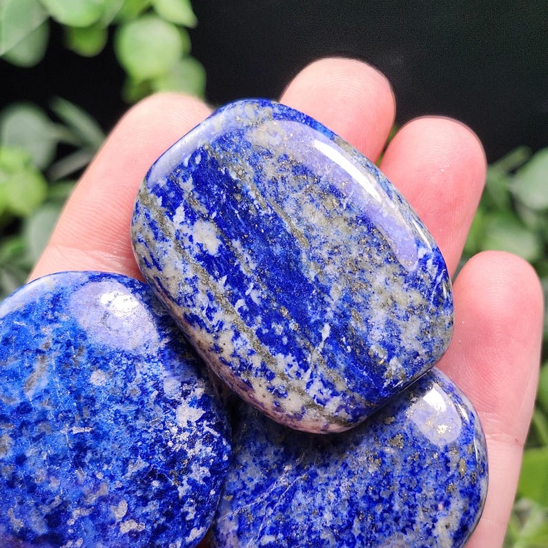 May include: A collection of polished lapis lazuli stones. The stones are a deep blue color with streaks of white and gold. The stones are smooth and rounded, and they are held in a hand.