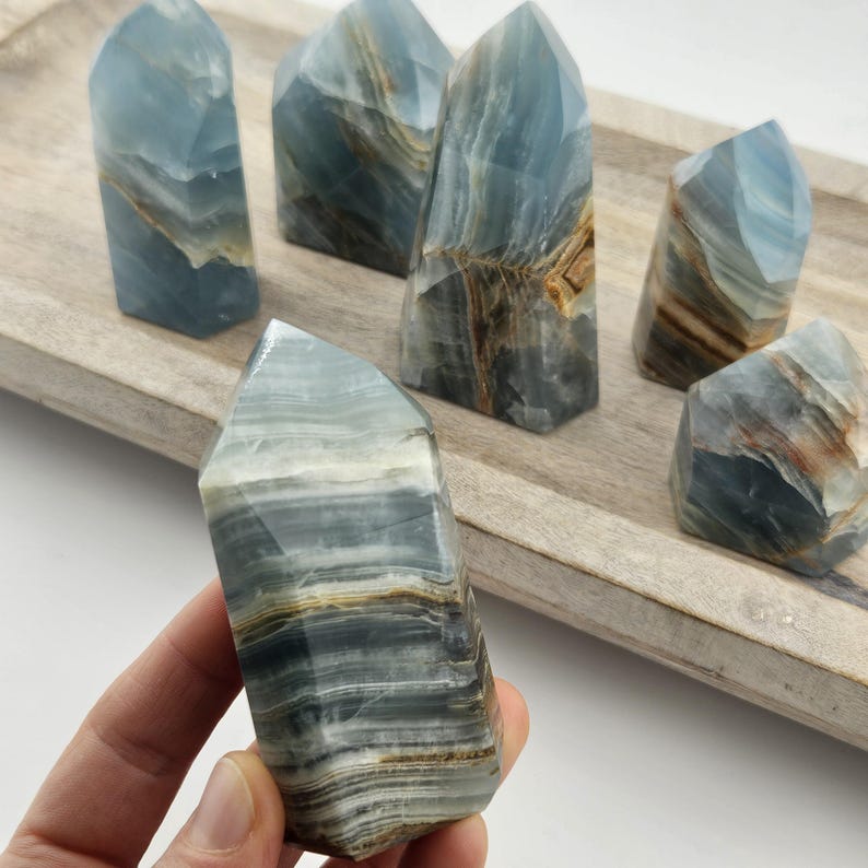 May include: Six blue onyx crystal towers with natural banding and varying shades of blue, brown, and white. One tower is held in a hand, showcasing the stone's polished surface and unique patterns. The crystals are displayed on a wooden tray.