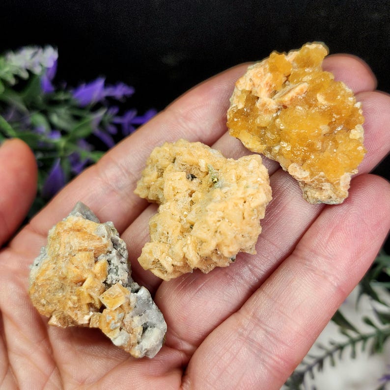 May include: Three raw, unpolished mineral specimens in shades of yellow, orange, and gray are displayed. The minerals have a rough, textured appearance, with visible crystalline structures. The background is blurred, with hints of purple and green.