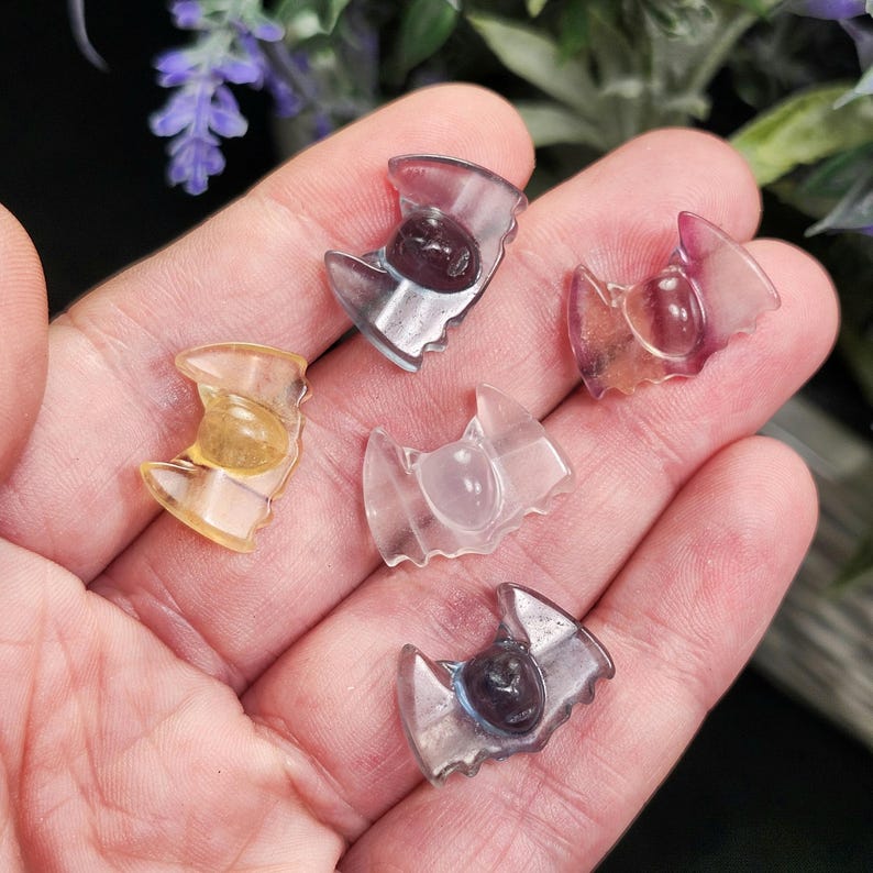 May include: Five small, translucent bat-shaped crystal carvings in shades of yellow, purple, and clear. Each bat has a rounded body and outstretched wings. The crystals are held in a hand against a blurred background of greenery.