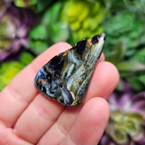 May include: A polished, teardrop-shaped piece of blue pietersite gemstone featuring swirling patterns of deep navy blue, bright sky blue, and metallic gold inclusions. The stone has a smooth, reflective surface with a distinct chatoyant luster.
