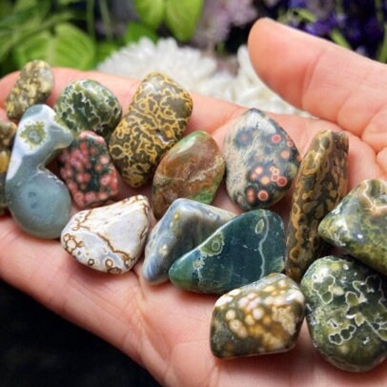 May include: A collection of polished, multi-colored, and patterned ocean jasper stones. The stones are various shapes and sizes, with shades of green, brown, white, and pink.