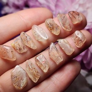 May include: A selection of clear, polished gemstones with internal inclusions. The stones are a light amber colour with dark brown and gold streaks. The gemstones are irregularly shaped and appear to be held in a hand.