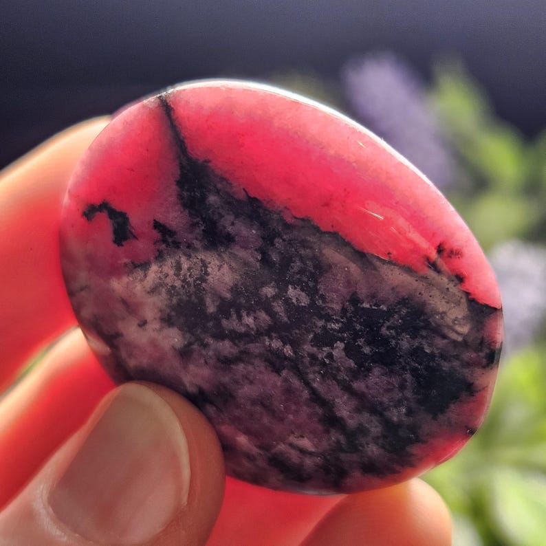 May include: A polished, circular rhodochrosite stone featuring a vibrant hot pink upper section and a dark, mottled black and grey lower section with intricate, vein-like patterns throughout the smooth, rounded surface of the mineral specimen.