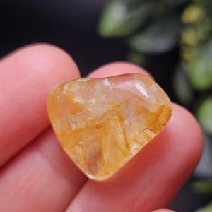 May include: A polished, heart-shaped golden rutilated quartz gemstone featuring translucent yellow tones and visible needle-like inclusions throughout the stone, showcasing a smooth, reflective surface and a distinct, rounded heart silhouette.