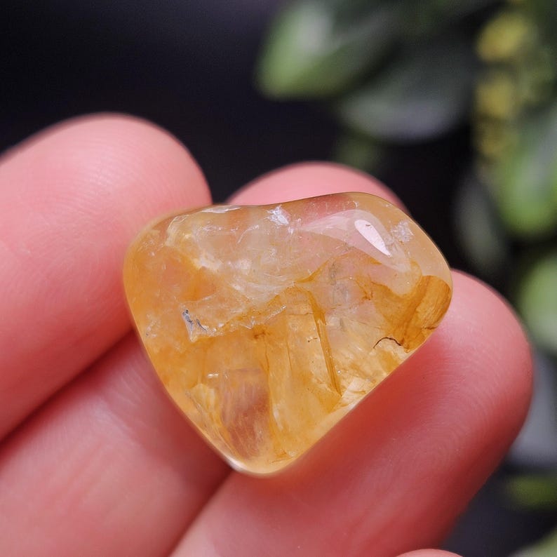 May include: A polished, heart-shaped golden rutilated quartz gemstone featuring translucent yellow tones and visible needle-like inclusions throughout the stone, showcasing a smooth, reflective surface and a distinct, rounded heart silhouette.