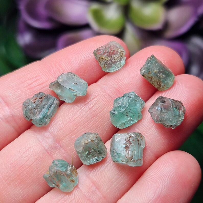 May include: A collection of nine small, rough, green gemstones. The gemstones are various shades of green and have a translucent appearance.