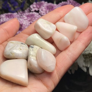 May include: A collection of smooth, polished tumbled stones in shades of pale pink, creamy white, and off-white. The stones vary in size and shape, featuring a glossy, reflective surface with subtle natural veining and occasional dark mineral inclusions.