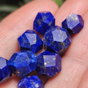 May include: A collection of faceted lapis lazuli gemstones with a deep blue color and visible gold-colored pyrite inclusions. Each stone features a geometric, multi-sided cut that creates sharp edges and reflective surfaces across the cluster.