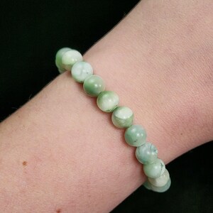 Peut inclure: Un bracelet compos&eacute; de perles rondes vert clair avec des marbrures blanches. Le bracelet est port&eacute; sur un poignet, sur fond sombre. Les perles mesurent environ 1 cm de diam&egrave;tre.