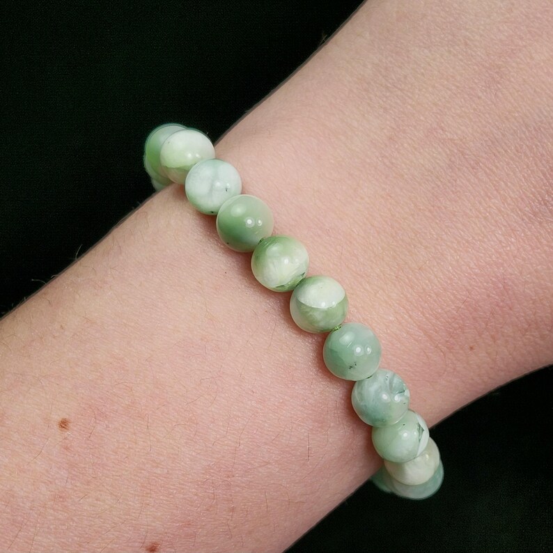 Peut inclure: Un bracelet compos&eacute; de perles rondes vert clair avec des marbrures blanches. Le bracelet est port&eacute; sur un poignet, sur fond sombre. Les perles mesurent environ 1 cm de diam&egrave;tre.
