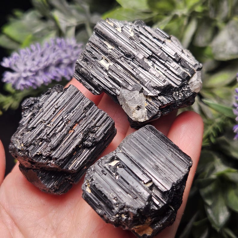 Peut inclure: Trois cristaux de tourmaline noire brute avec des stries verticales distinctes et une surface brillante et r&eacute;fl&eacute;chissante. Les pierres pr&eacute;sentent des bords dentel&eacute;s et irr&eacute;guliers ainsi que de petites inclusions min&eacute;rales claires.