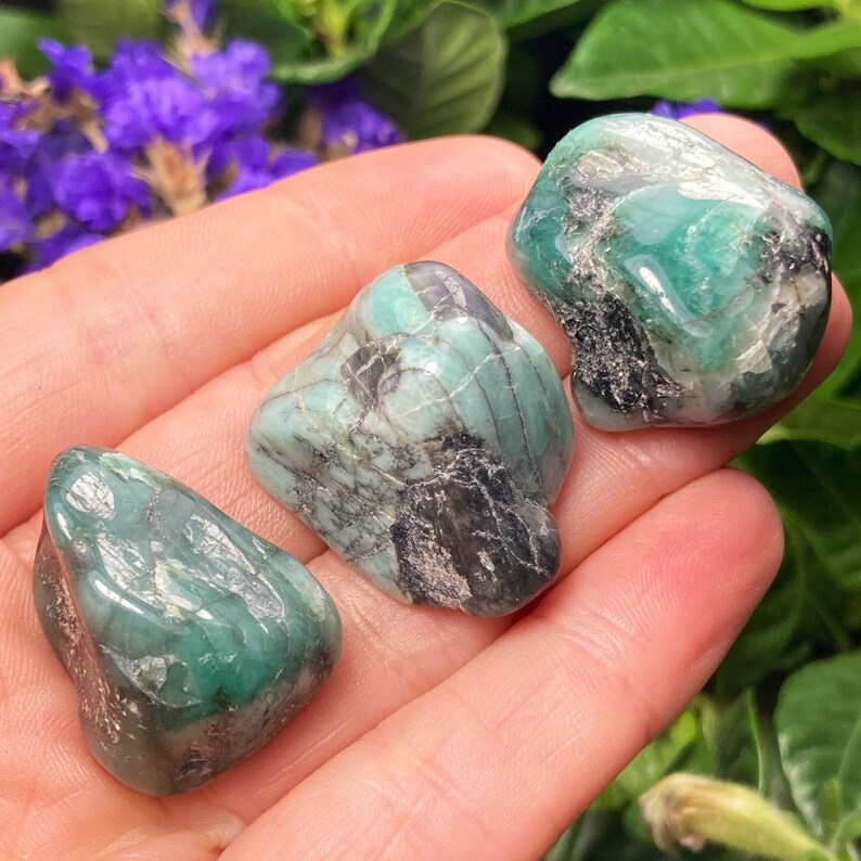 May include: Three polished emerald gemstones. The stones are a deep green colour with black inclusions and a smooth, rounded shape. The gemstones are displayed against a soft background of a hand and green foliage.