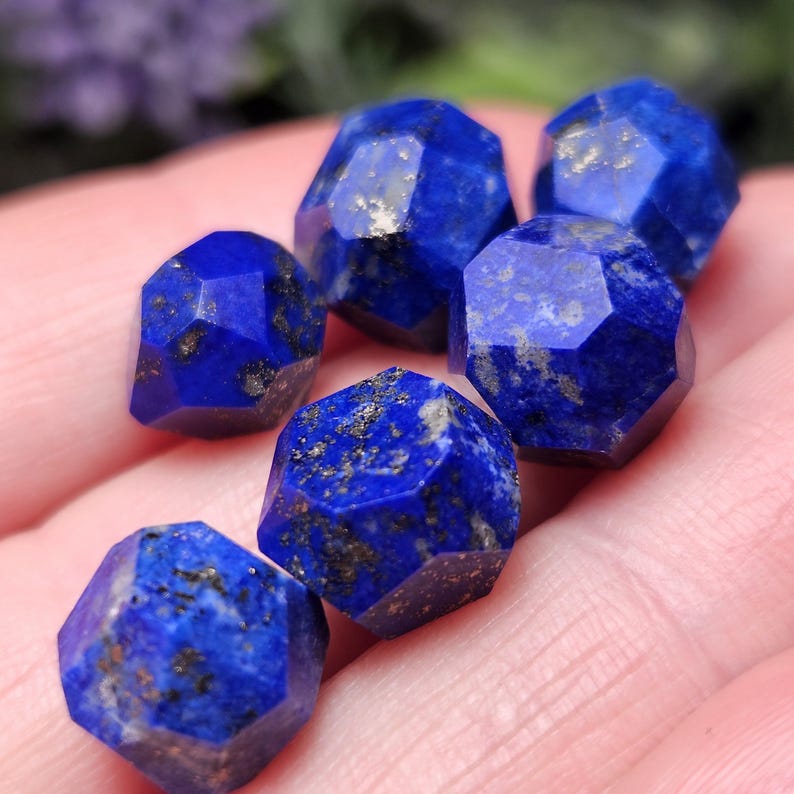 May include: A collection of six small, faceted lapis lazuli gemstones. Each stone features a deep blue color with visible gold-colored pyrite inclusions. The stones have a geometric, multi-sided cut with sharp edges and a polished, reflective surface.
