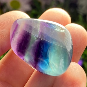 May include: A polished, oval-shaped fluorite stone. The stone displays bands of purple, blue, and green hues. The translucent gemstone is held in a hand, with a blurred green background.
