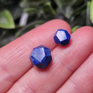 May include: Two faceted lapis lazuli gemstones with a deep blue color and a geometric, multi-sided cut. The stones are polished to a smooth finish, showcasing the natural texture and rich, saturated blue tones of the mineral in a small size.