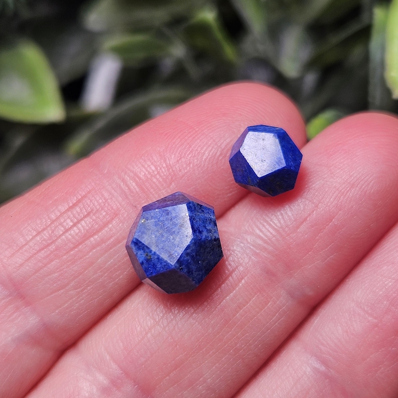 May include: Two faceted lapis lazuli gemstones with a deep blue color and a geometric, multi-sided cut. The stones are polished to a smooth finish, showcasing the natural texture and rich, saturated blue tones of the mineral in a small size.