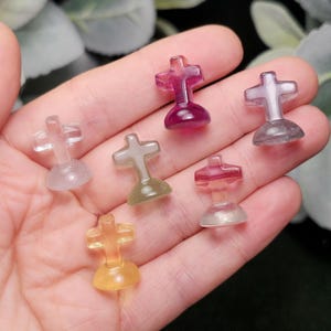 May include: A collection of small, translucent cross-shaped figurines in various colors, including purple, yellow, and clear. The crosses are held in a hand, showcasing their polished surfaces and rounded bases. The figurines are likely made of a gemstone or crystal.