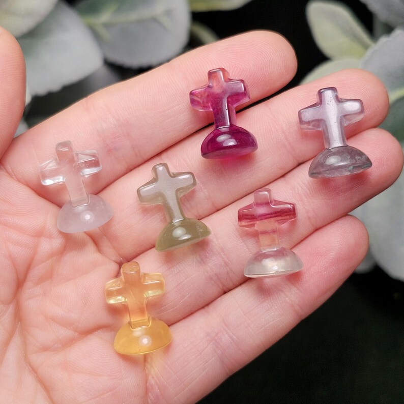 May include: A collection of small, translucent cross-shaped figurines in various colors, including purple, yellow, and clear. The crosses are held in a hand, showcasing their polished surfaces and rounded bases. The figurines are likely made of a gemstone or crystal.
