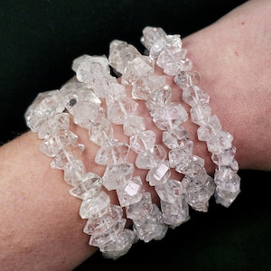 Könnte beinhalten: Armbänder aus klarem Quarz-Kristall. Die Armbänder bestehen aus unregelmäßigen, facettierten Quarz-Kristallen, die aufgefädelt sind. Die Kristalle sind durchscheinend und reflektieren das Licht. Die Armbänder werden am Handgelenk getragen.