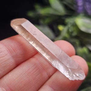 May include: A long, slender, clear quartz crystal point with a smooth, faceted surface and a subtle reddish-brown tint near the top. The crystal has a pointed termination and a slightly jagged, uneven base, showcasing natural internal inclusions.