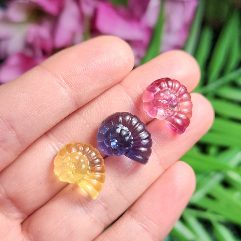 Peut inclure: Trois petites sculptures de pierres pr&eacute;cieuses translucides en forme de spirale, dans les tons jaune, violet et rose. Les sculptures sont tenues dans une main, avec un arri&egrave;re-plan flou de fleurs roses et de feuillage vert.