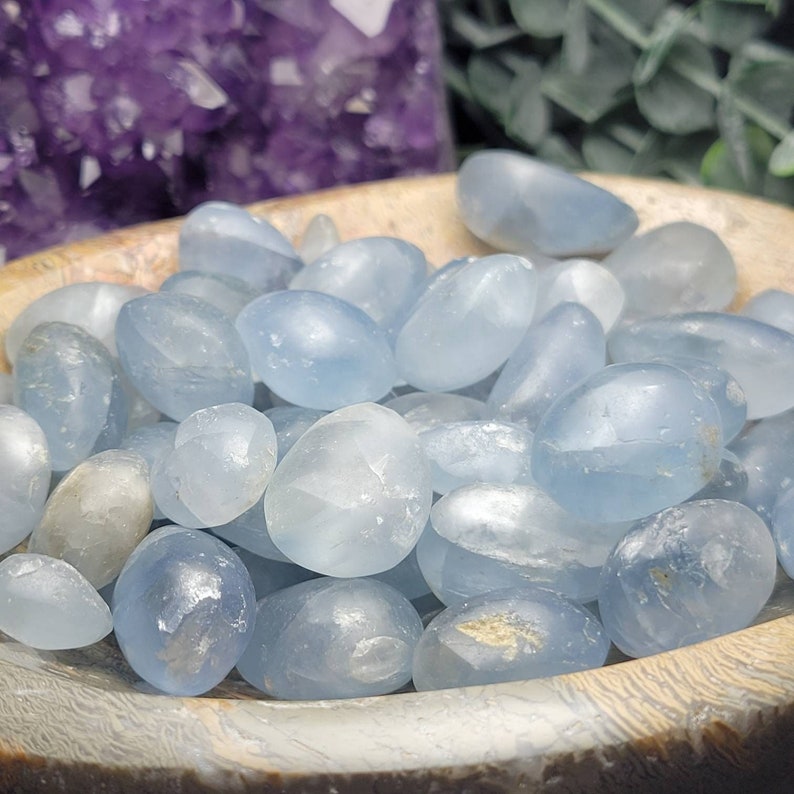 May include: A bowl filled with light blue tumbled celestite stones. The stones are smooth and polished, with a slightly cloudy appearance.