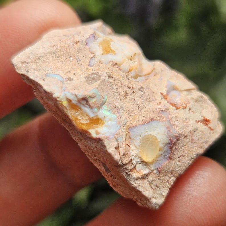 May include: A rough, light tan rock specimen featuring embedded patches of iridescent opal. The stone displays natural inclusions with translucent yellow and white mineral deposits that exhibit flashes of green and blue light across the surface.