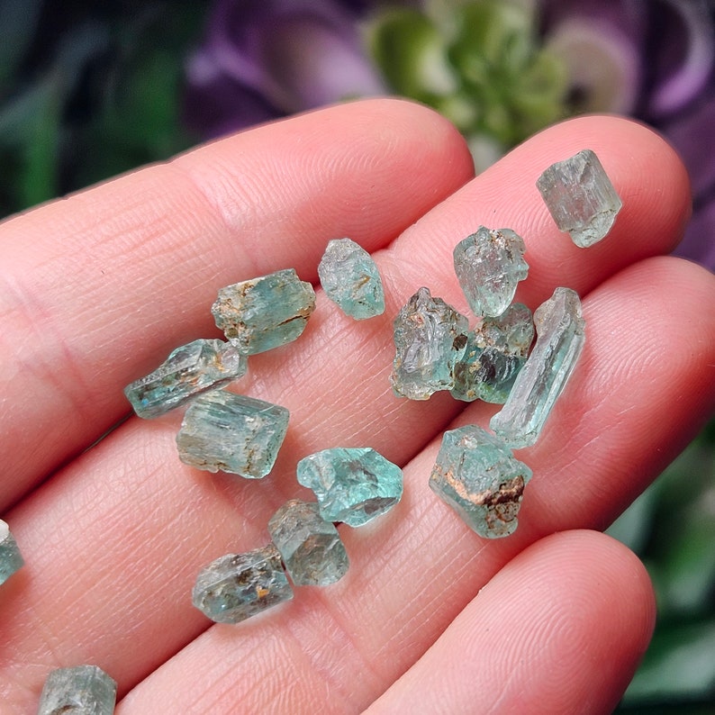 May include: A collection of small, light blue and green gemstones, likely emeralds, displayed on a hand. The gemstones are rough and have a natural, unpolished look.