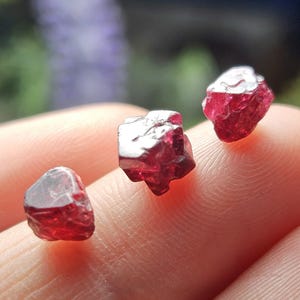 May include: Three red ruby crystals, each with a different shape and size, are displayed on a finger.