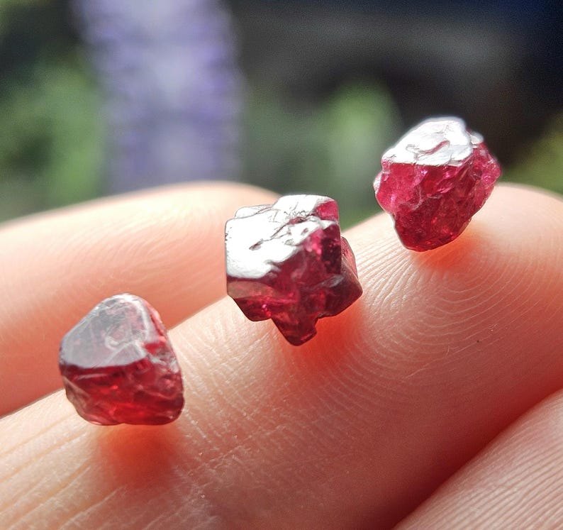 May include: Three red ruby crystals, each with a different shape and size, are displayed on a finger.