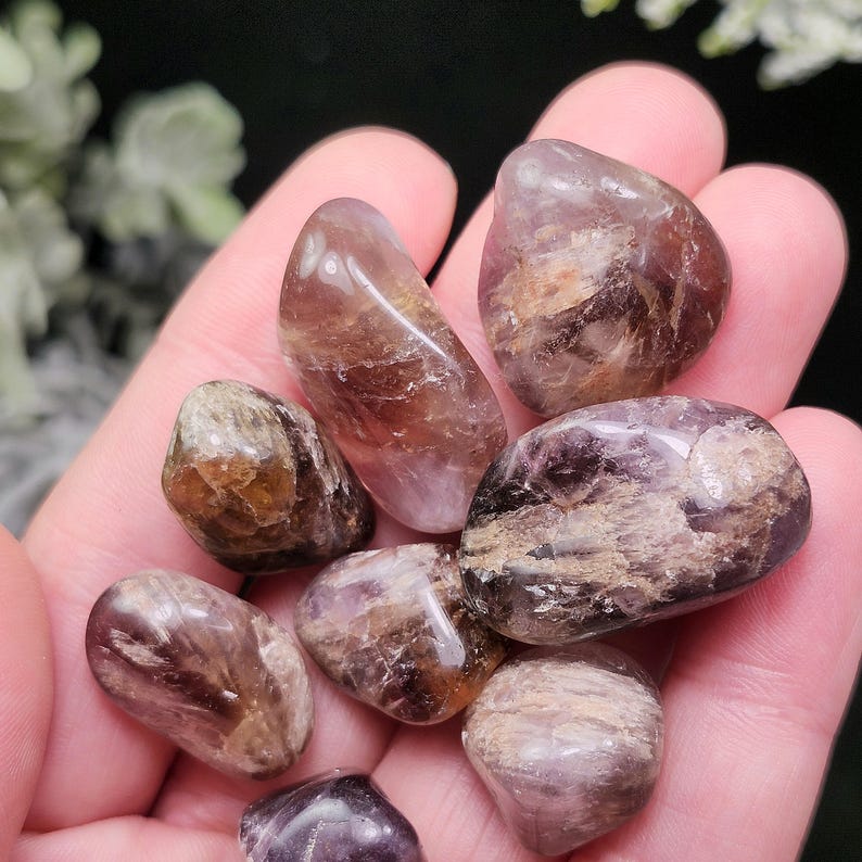 May include: A collection of tumbled amethyst stones in various shades of purple, brown, and white. The stones are smooth and polished, with natural patterns and textures. The stones are held in a hand.