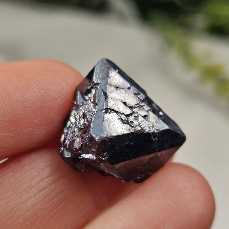 Puede incluir: Un cristal oscuro y facetado con un brillo met&aacute;lico p&uacute;rpura oscuro y plateado. La superficie del cristal tiene una apariencia texturizada y fracturada, captando la luz y creando un efecto brillante. El cristal se sostiene en una mano.