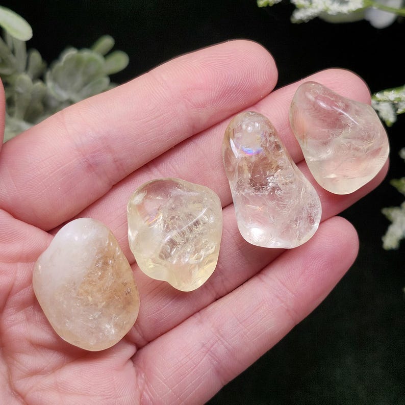 May include: Four polished, tumbled citrine gemstones in varying shapes and sizes. The stones range in color from pale yellow to a golden hue, with a translucent quality. The gemstones are held in a hand, with a blurred green background.