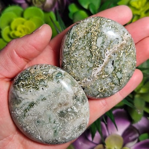 May include: Two polished, round stones with a mottled pattern of green, white, and gold. The stones are held in a hand, showcasing their smooth, reflective surfaces. The natural stones have a unique, organic appearance.