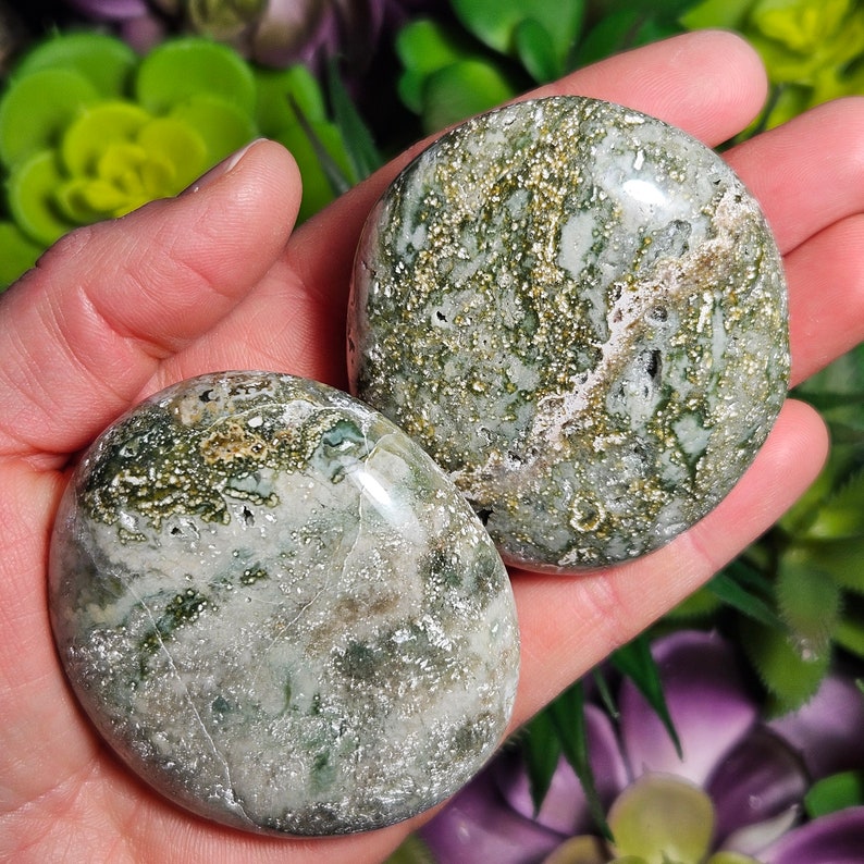 May include: Two polished, round stones with a mottled pattern of green, white, and gold. The stones are held in a hand, showcasing their smooth, reflective surfaces. The natural stones have a unique, organic appearance.