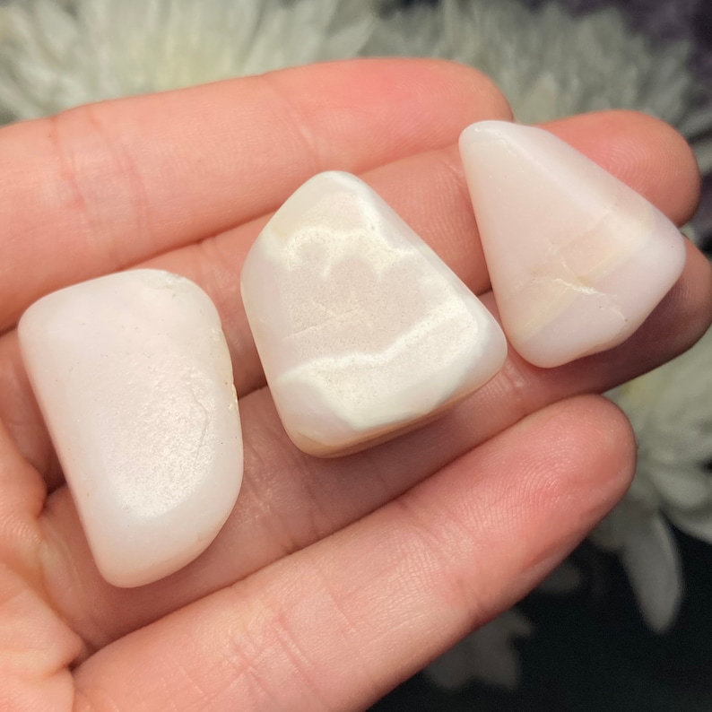 May include: Three polished, tumbled stones with a smooth, opaque, pale pink to off-white appearance. Each stone features a rounded, irregular shape with subtle variations in colour density and a soft, reflective surface texture.