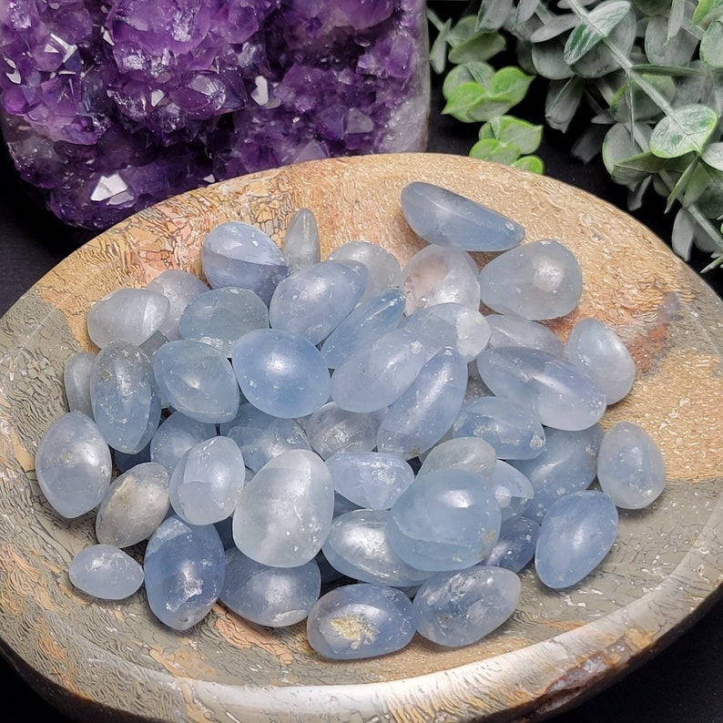 May include: A bowl filled with light blue tumbled celestite stones. The stones are smooth and polished, with a translucent appearance.