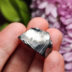 May include: A dark gray, metallic, irregularly shaped rock with a striated surface. The rock has a silvery sheen and appears to be a piece of meteorite.