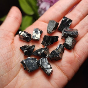 May include: A collection of black obsidian crystal chips, with sharp edges and a shiny, reflective surface. The chips are arranged on a hand, showcasing their natural, raw form.