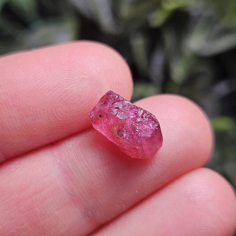 May include: A small, raw, unpolished gemstone with a vibrant, translucent hot pink colour. The stone features an irregular, multifaceted surface with natural textures and internal inclusions, showcasing a deep magenta hue throughout its structure.