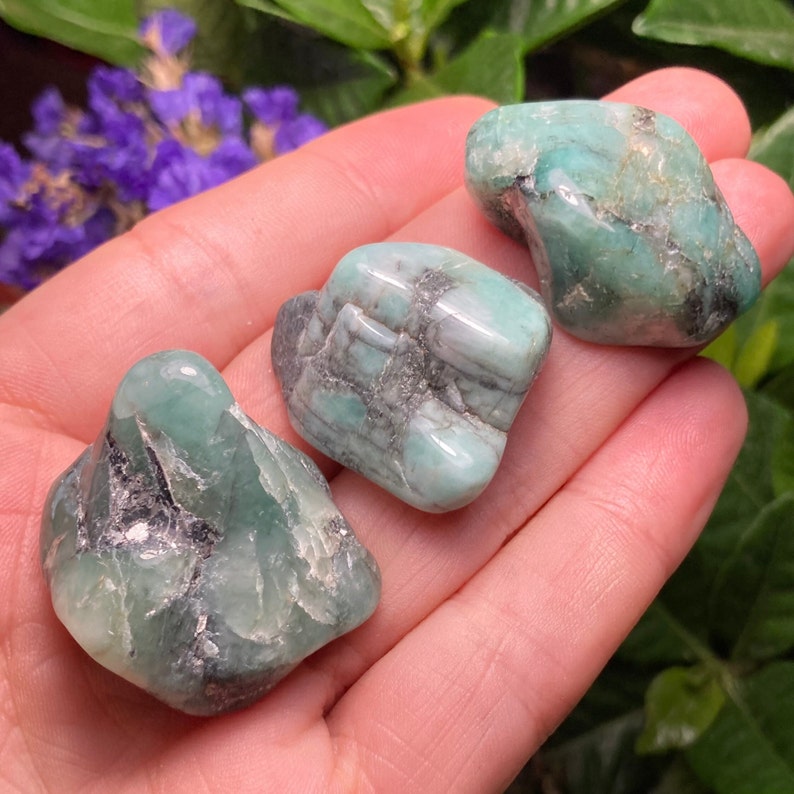 May include: Three polished emerald gemstones, each with a unique shape and a vibrant green hue. The stones have natural inclusions and a slightly translucent appearance, resting in a hand.