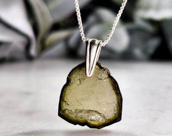 May include: A silver necklace featuring a pendant made from a rough-cut, olive-green gemstone. The pendant is irregularly shaped with a silver bail, suspended from a delicate silver chain. The background is blurred, with hints of green foliage.