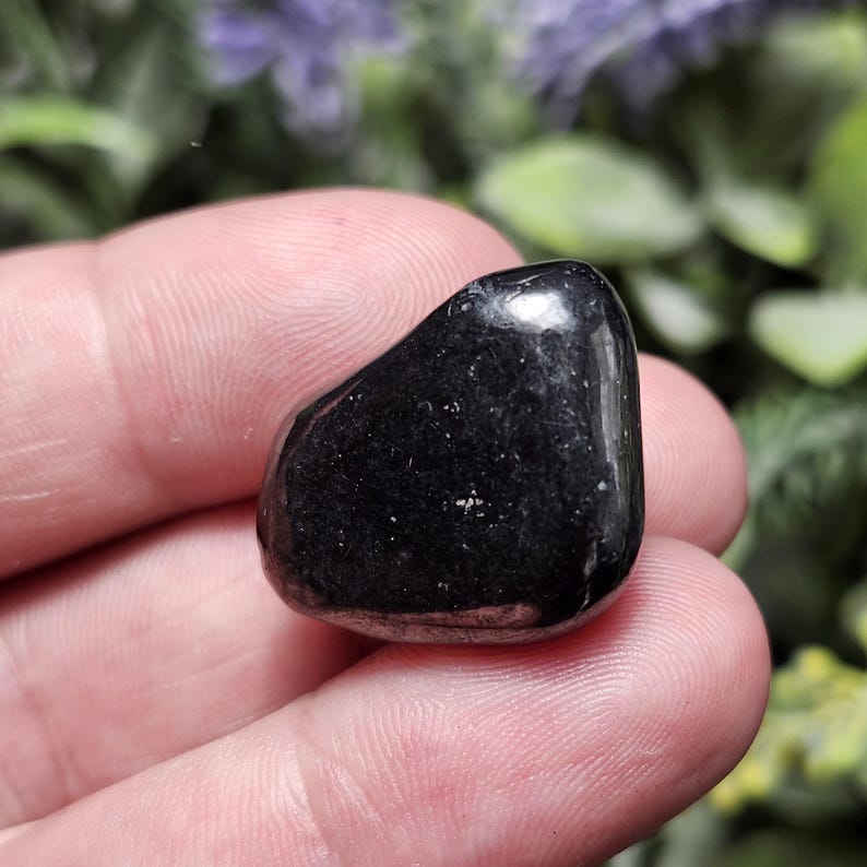 May include: A smooth, black, polished stone with a slightly rounded shape. The stone is held in a hand.