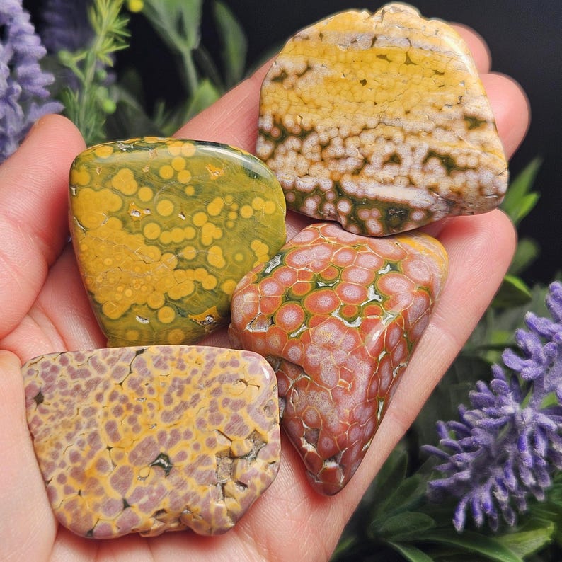 May include: A hand holds four polished gemstones. The stones display a variety of colors, including yellow, green, red, and purple, with unique patterns. The gemstones are irregularly shaped and appear to be natural specimens, possibly for collection or display.