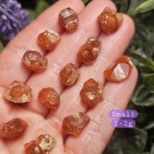 May include: A collection of small, raw, orange-brown crystals. The crystals vary in shape and size, with a rough, natural texture. The image includes text that reads "Small 1-2g". The crystals are displayed against a blurred background of green and purple.
