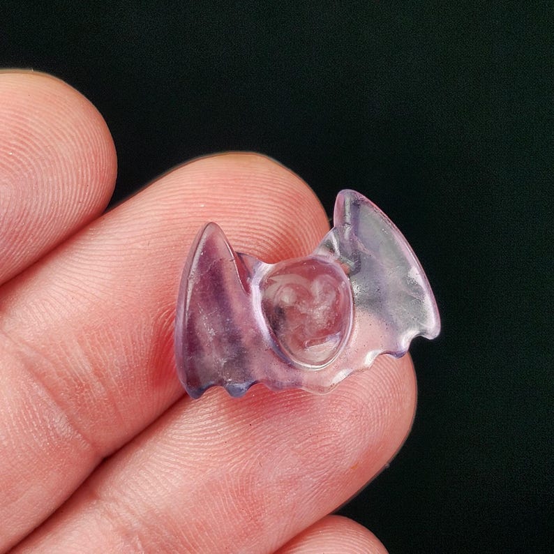 May include: A small, translucent purple bat-shaped carving. The carving has a heart-shaped center and wing-like extensions. The object is held between fingers, with a dark green background.