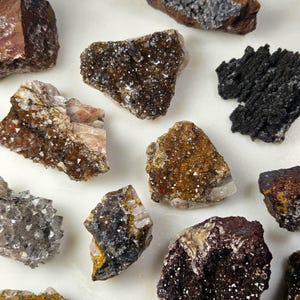 May include: A collection of various raw mineral specimens, including quartz and other crystalline formations. The stones display a range of colors, from deep browns and blacks to lighter tans and grays, with sparkling crystal inclusions.